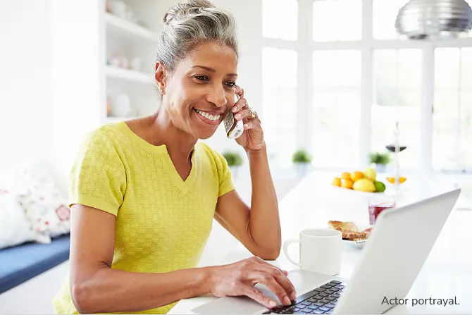 Actor portrayal of a woman with gray hair in a yellow shirt sitting at her counter on the phone while looking at her laptop